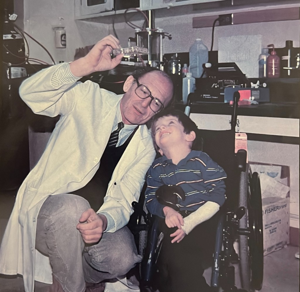 Dr. Baylink in his laboratory holding a culture plate up to the light beside a boy in a wheelchair